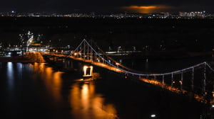 Night shot of a pedestrian bridge in Kyiv over the Dnieper, illuminated by warm lights that light up the surroundings.