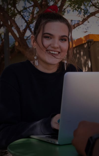 A woman with a beaming smile sits at a table and works on her laptop